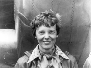 black and white photo of Amelia Earhart standing under the nose of her Lockheed Model 10 plane, it's a close-up shot with her smiling, wearing large collared shirt and scarf tied around the neck