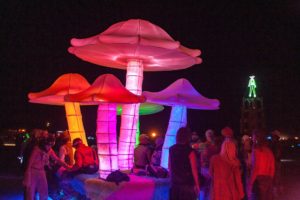 five giant mushroom lit in a rainbow of colors, surrounded by people, the scene is taken at night