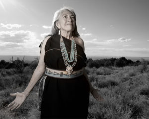 older woman dressed in Native American attire with beaded necklace facing camera, looking at the sun, arms outstretched, photo is black and white, background is open sky and sagebrush-covered ground