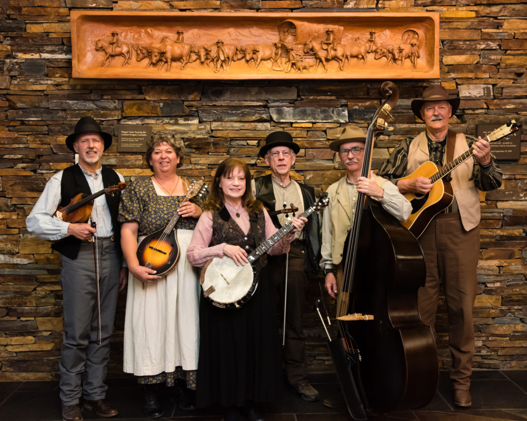 six people standing in front of an indoor rock wall all hold instruments and dressed in turn-of-the-century clothing
