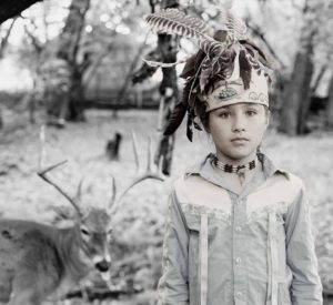 black and white photo of young boy (8 years old) in western denim shirt and Native American headdress standing and looking at camera, serious, in the background on the left side is a deer with big antlers sitting on the ground, looking towards the boy